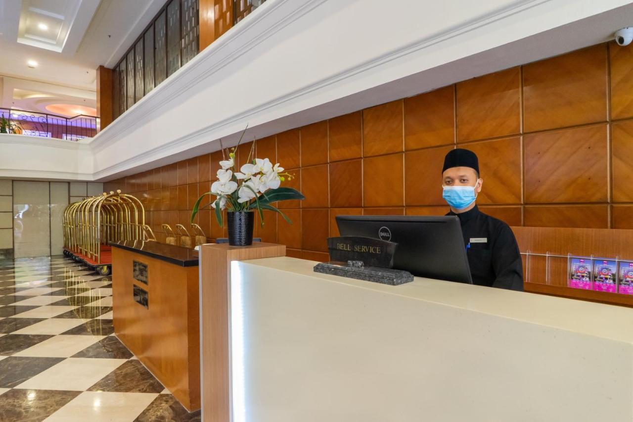 Seri Pacific Hotel Kuala Lumpur Exterior photo The photo shows a hotel reception area. In the foreground, a staff member is standing behind a reception desk, wearing a mask and a dark uniform. The desk has a computer monitor, and there are decorative elements, including a vase with flowers. Behin