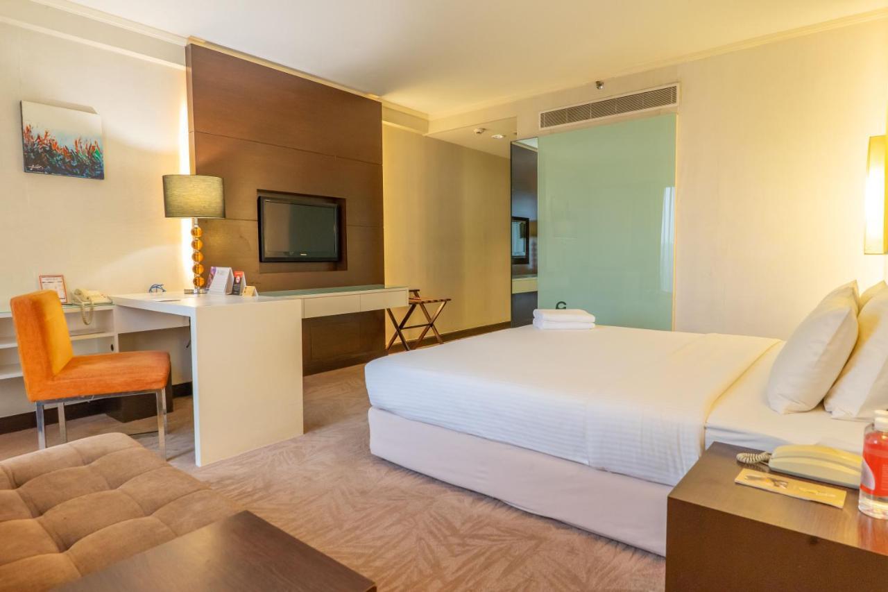 Seri Pacific Hotel Kuala Lumpur Exterior photo The photo shows a hotel room or a similar accommodation space. It features a large bed with white bedding, a bedside table with a lamp, and a small seating area. There is a desk with a chair, and a television mounted on the wall. A glass partition se