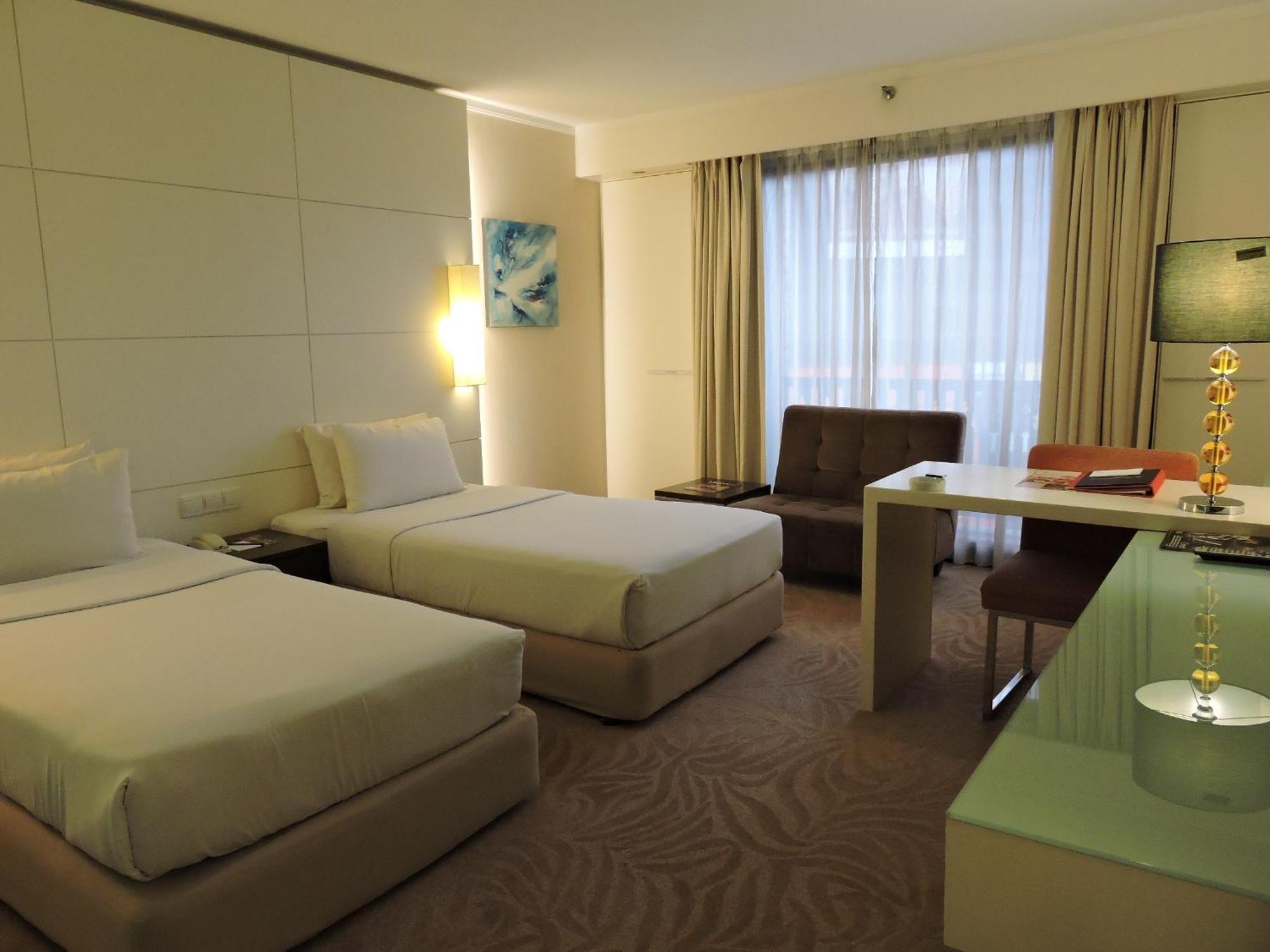 Seri Pacific Hotel Kuala Lumpur Room photo The photo shows a modern hotel room featuring two twin beds with white bedding. The walls are light-colored with a sleek and minimalist design. There is a large window covered by sheer curtains, allowing natural light to enter the room. To one side, 