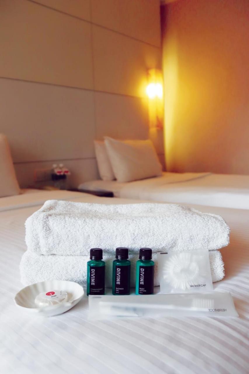 Seri Pacific Hotel Kuala Lumpur Exterior photo The photo shows a neatly arranged stack of white towels placed on a bed. In front of the towels, there are three small green bottles, likely containing shampoo, conditioner, and body wash. The background features a well-decorated hotel room with two 